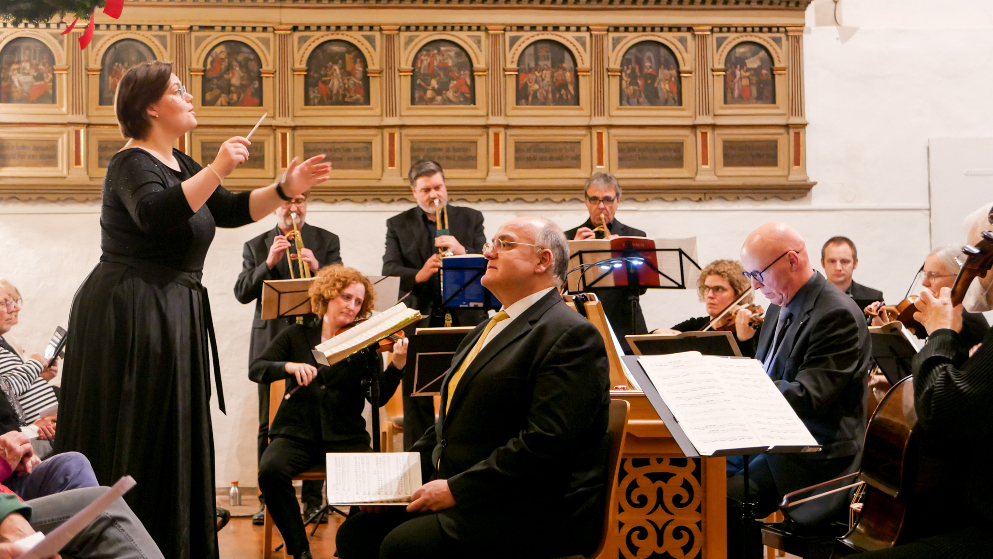 Ein Orchester mit einem Orgelpositiv in einer Kirche. Links eine Dirigentin.