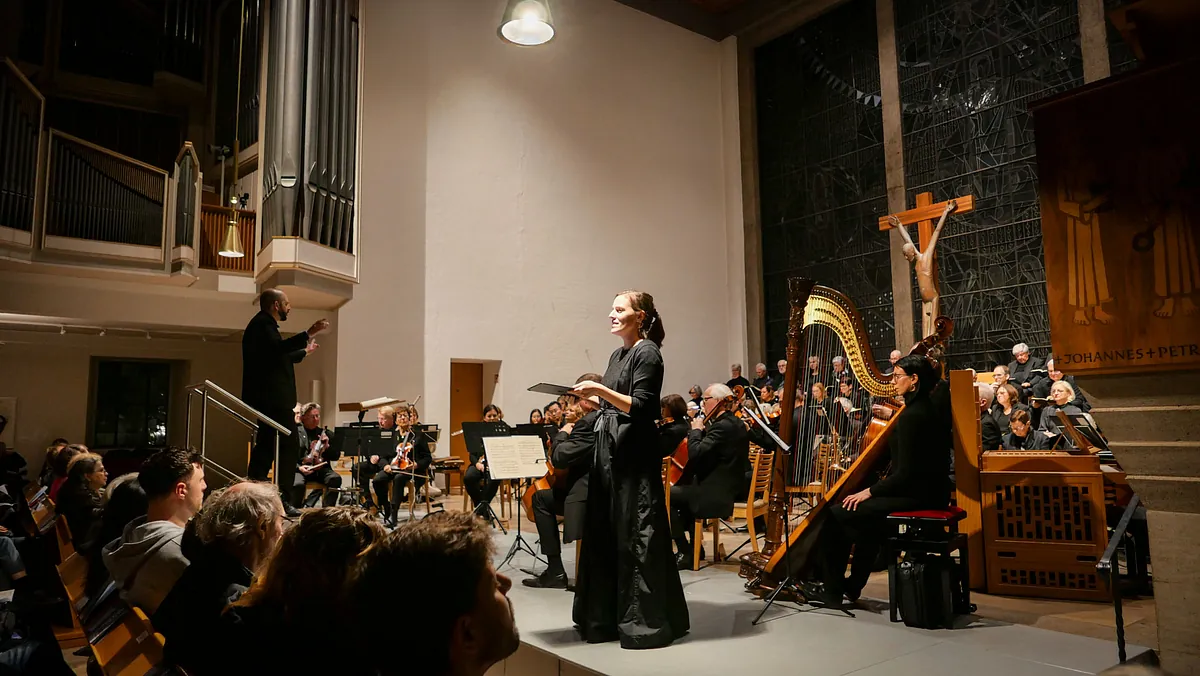 Eine Solisten im schwarzen Kleid singt in einer Kirche. Hinter ihr ein Orchester und sitzend ein gemischter Chor