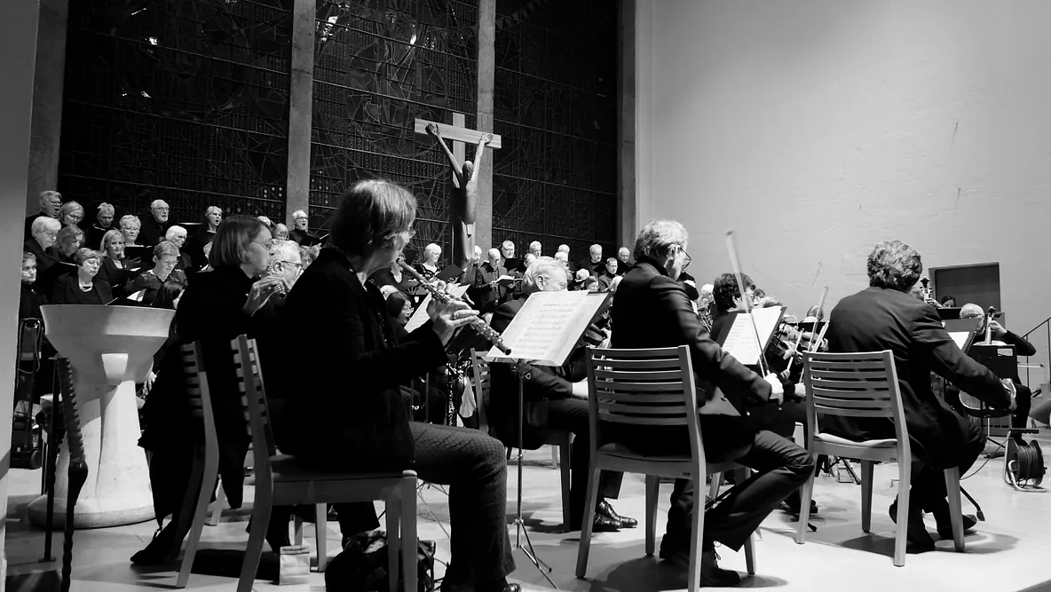 Ein Orchester von der Seite, über die Schultern von Flötenspielerinnen gesehen. Im Hintergrund ein Chor vor einem modernen Kruzifix