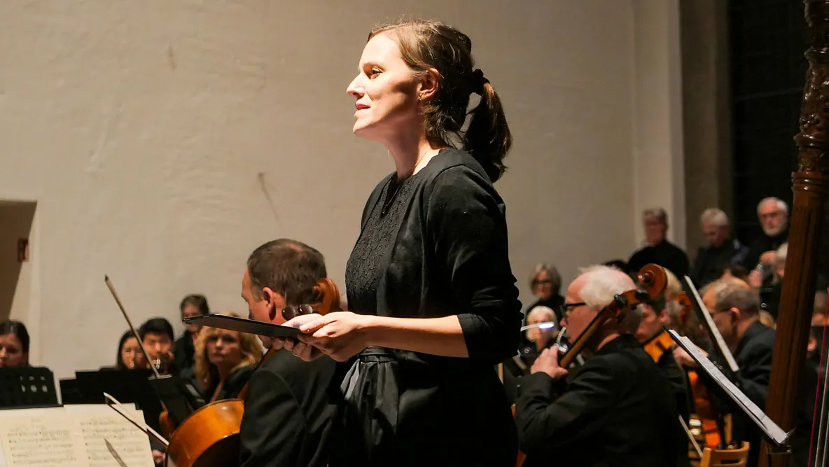 Eine Solistin im schwarzen Kleid in Nahaufnahme. Im Hintergrund Mitglieder eines Orchesters und Chors