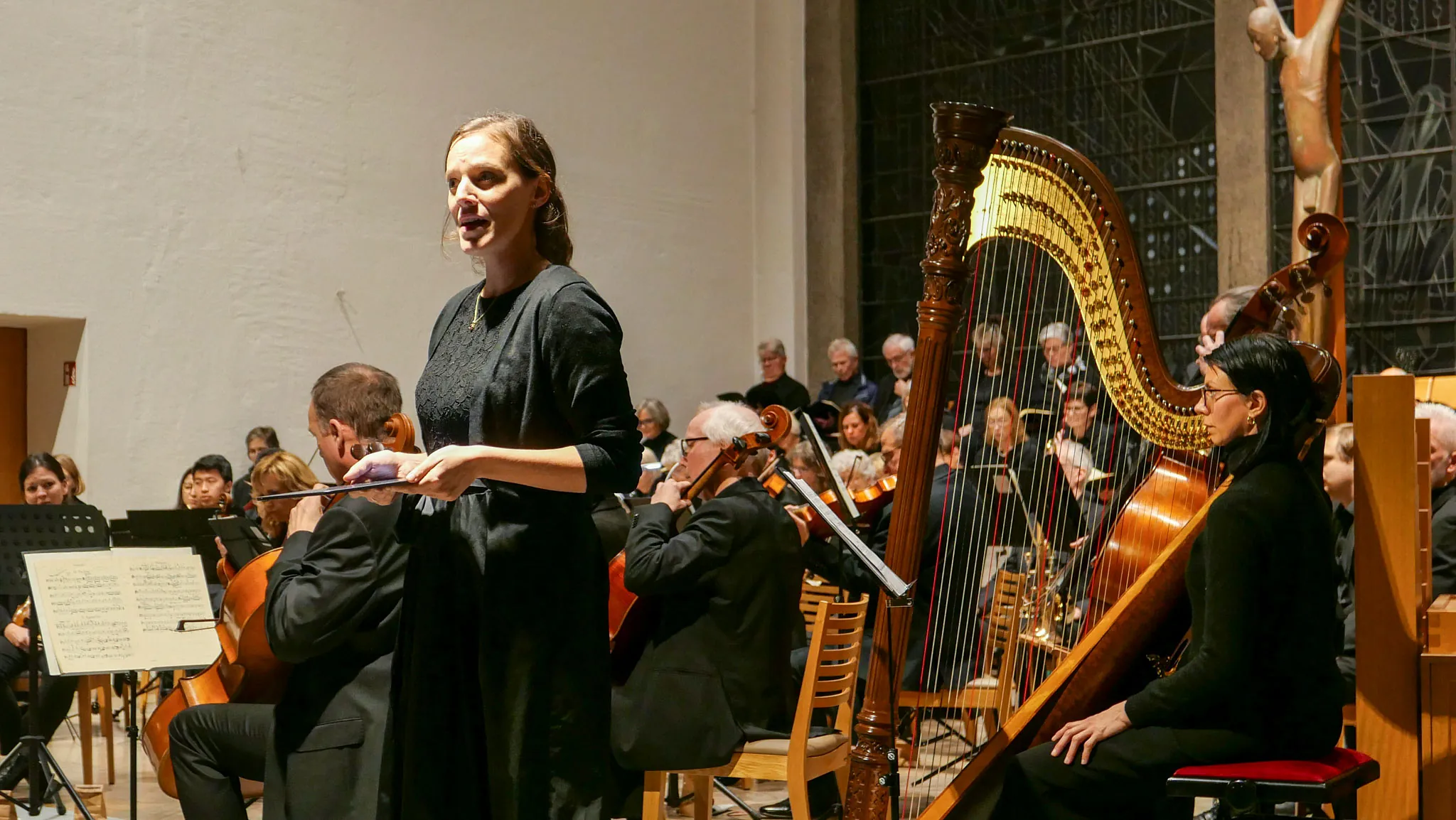 Eine Solistin singt in einer Kirche. Neben ihr eine Harfe. Im Hintergrund ein Orchester und ein sitzender Chor