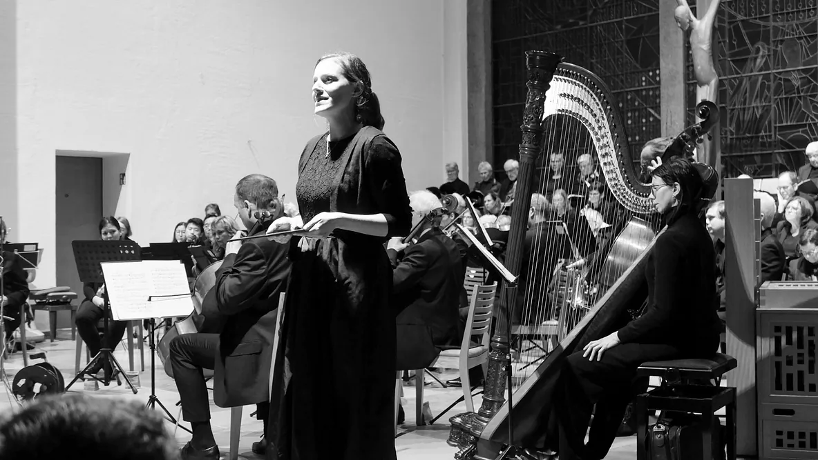 Ein Schwarzweiß-Bild einer Solistin, die vor einem Orchester und einem Chor in einer Kirche singt
