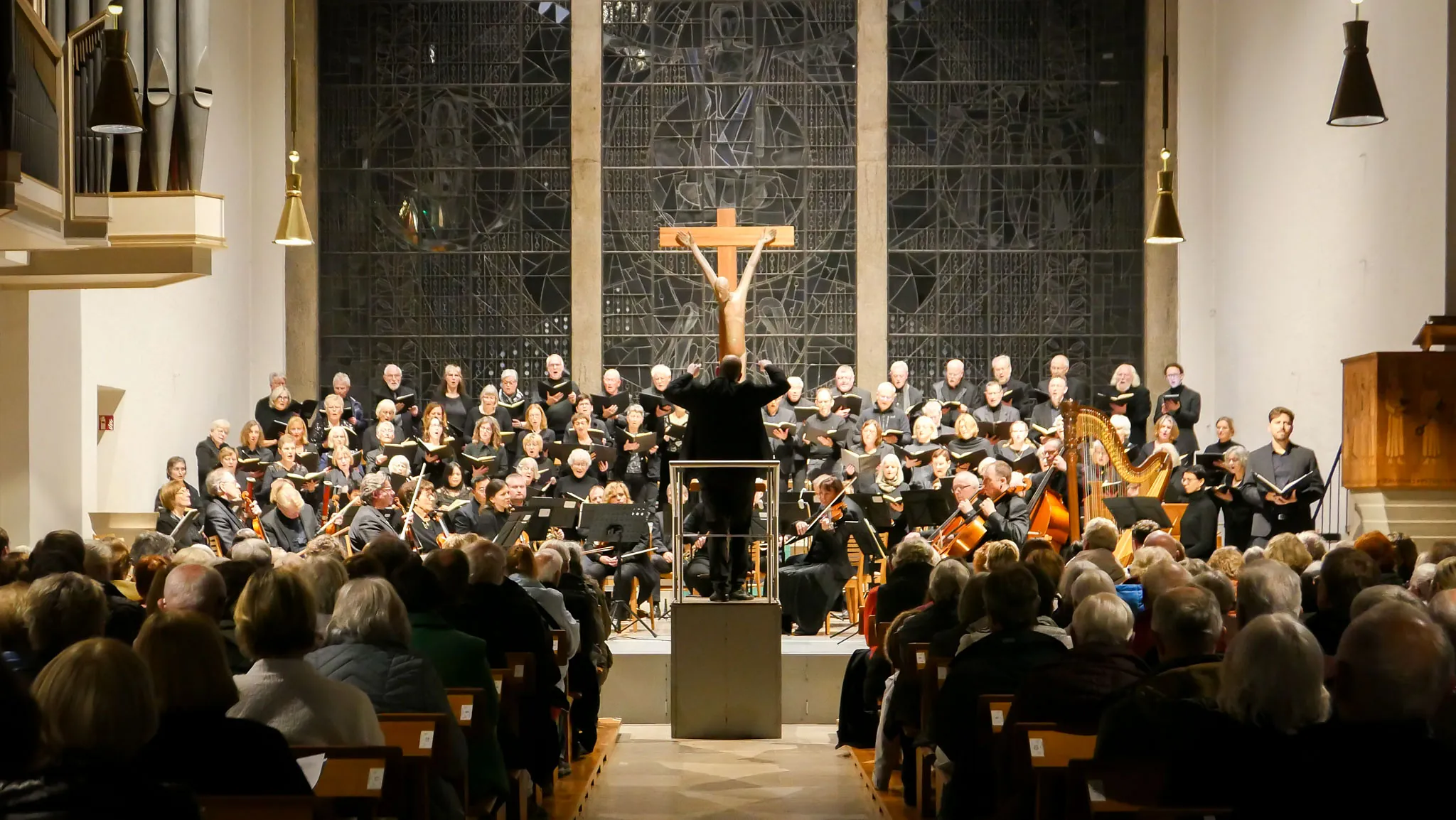 Ein Dirigent leitet ein Orchester und einen Chor in einer Kirche vor einem großen dunklen Kirchenfenster und einem modernen Kruzifix. Im Vordergrund im Schatten das Publikum