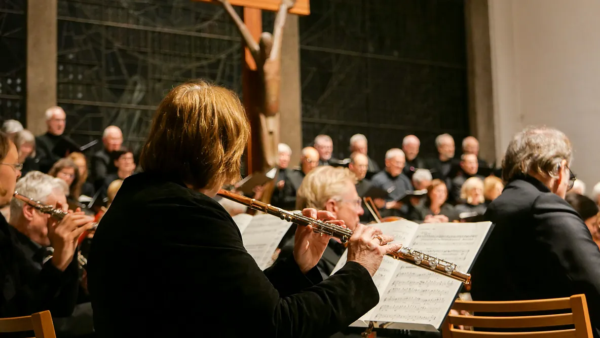 Eine Querflötenspielerin von hinten gesehen. Über ihre Schultern sieht man verschwommen weitere Mitglieder des Orchesters und Chors