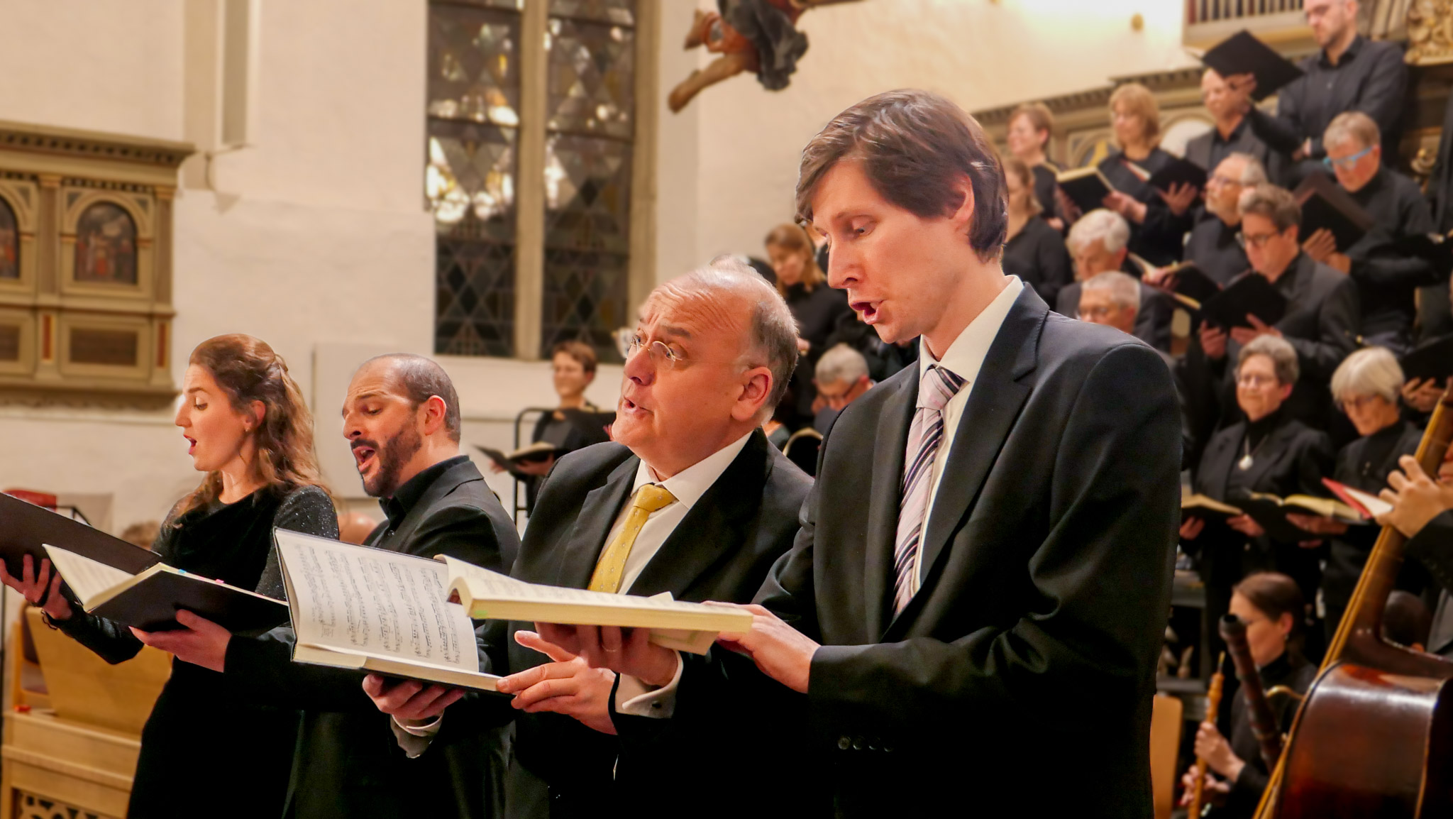 Eine Sängerin und drei Sänger in einer Kirche. Im Hintergrund ein gemischter Chor in Schwarz.